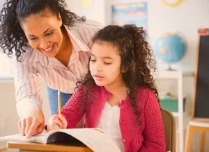A woman smiles and helps a young girl with curly hair as she writes in a notebook at a desk in a classroom.