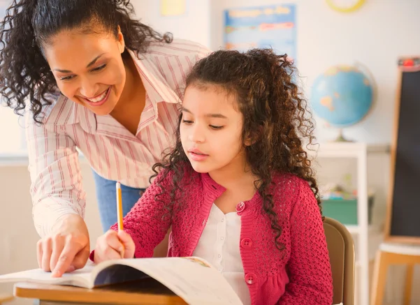 A teacher smiles and points while helping a young girl with her schoolwork at a desk in a classroom.