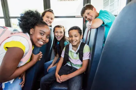 Five smiling children with backpacks sit together on a school bus, looking at the camera and appearing happy.