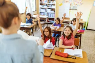 Children in a classroom raise their hands as a teacher stands at the front, holding a book and facing the students.