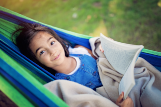 A young child looks up from a book while lounging in a hammock