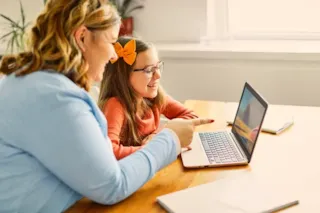 An adult and a child smile while looking at a laptop together at a wooden table in a bright room.