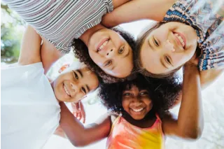 Four smiling children standing in a circle, arms around each other, looking down at the camera outdoors.