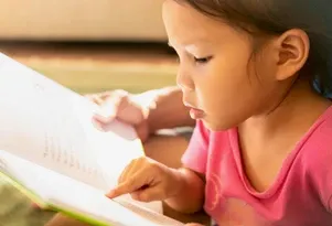 Young child in a pink shirt intently reading a book, pointing at the words with a finger in natural light.