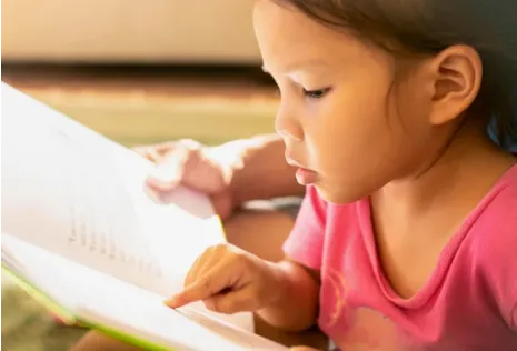 A young girl in a pink shirt reads a book, pointing at the words with her finger.