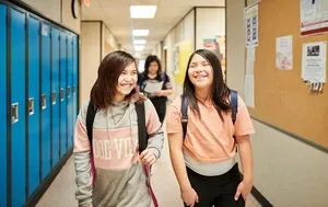 Two girls with backpacks smile and walk down a school hallway lined with lockers and bulletin boards.
