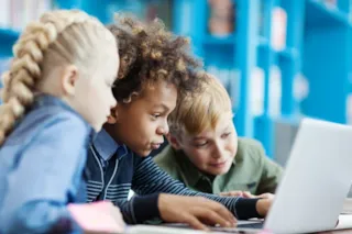 Three children sit close together, looking intently at a laptop screen in a brightly lit room with blue shelves.