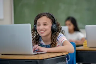 Smiling girl wearing headphones and using a laptop in a classroom with a chalkboard and another student in the background.