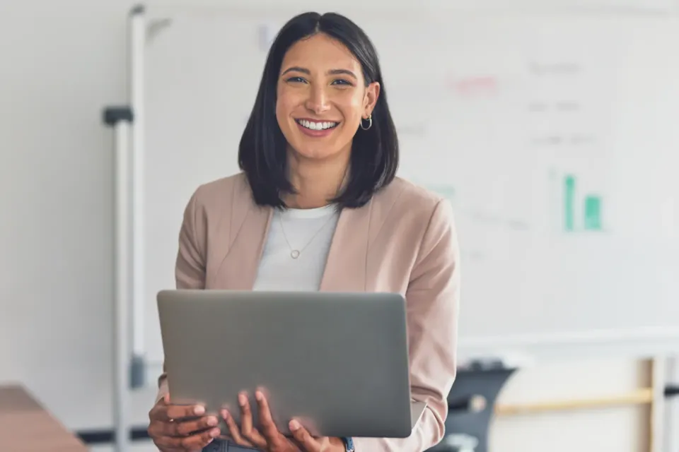 Woman smiling and holding a laptop, standing in front of a whiteboard with charts in an office setting.