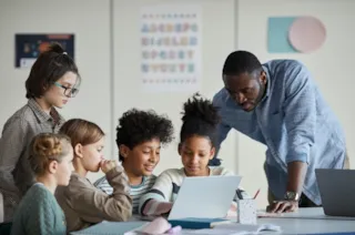A teacher helps a group of diverse students working together on a laptop in a classroom.