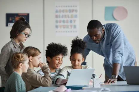 A teacher helps a group of smiling students working together at a desk with a laptop in a classroom.