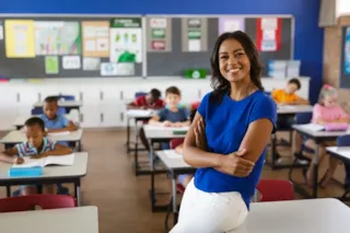 A teacher smiles confidently in a classroom with young students seated at desks and engaged in their work.