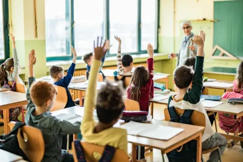 Students in a classroom raising their hands while a teacher stands at the front, engaging with the class.