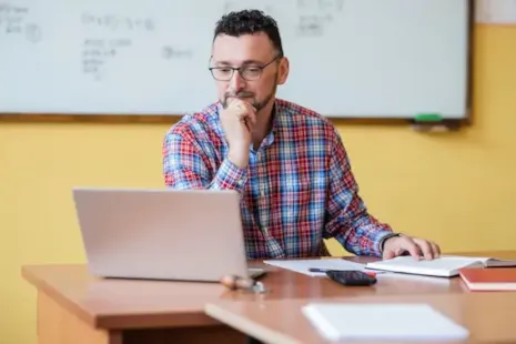 Man in glasses and plaid shirt works on a laptop at a desk, with papers and a whiteboard in the background.