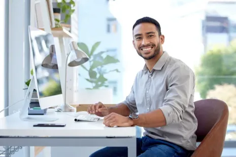 Smiling man sitting at a desk with a computer in a bright, modern office space.