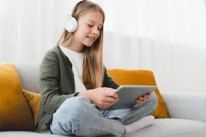 Girl wearing headphones sits cross-legged on a couch, smiling while using a tablet.