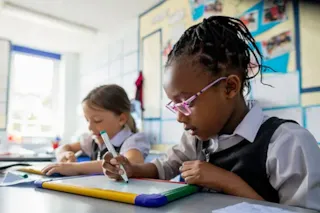 Two young girls in school uniforms write on whiteboards at their desks in a bright classroom.