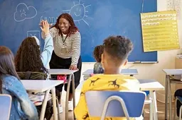 A teacher in the front of her classroom engages with her students, who are seated at their desks, raising their hands