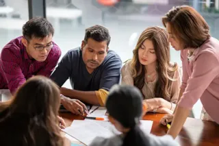 Five people gathered around a table, discussing documents and collaborating on a project in an office setting.