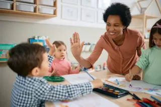 Teacher giving a high-five to a young boy as children paint at a classroom table.