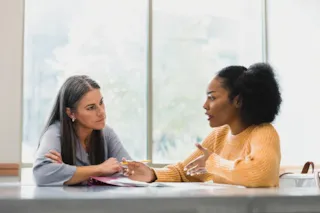 Two women sit at a table, having a serious conversation, with papers and pens in front of them.