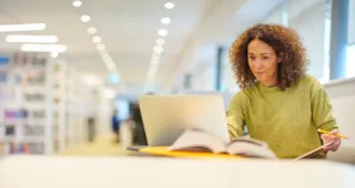 Woman with curly hair studying at a laptop in a bright library, with open book and pencil in hand.