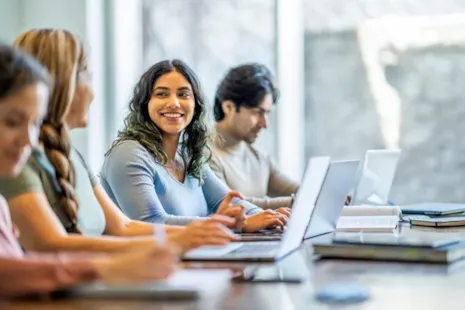 Four students sit at a table with laptops and books. One woman in the center smiles at another student.