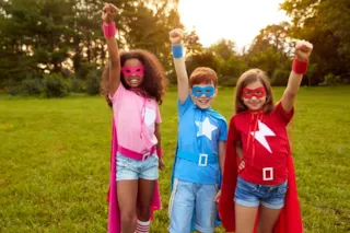 Three children wearing superhero costumes and masks stand outside with one arm raised, smiling confidently.