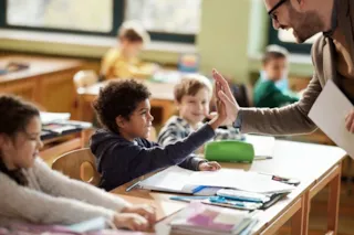 Smiling teacher gives a high-five to a student in a lively classroom with children at their desks.