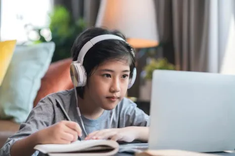 A child with headphones studies at a laptop, writing in a notebook at a desk in a cozy room.
