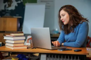 Woman in a blue sweater studies at a desk with a laptop and a stack of books in a classroom setting.