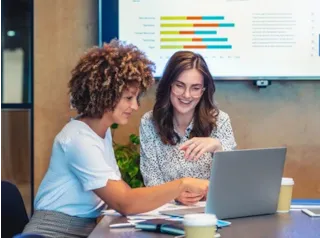 Two women smiling and discussing data on a laptop in an office, with a large presentation screen displaying colorful charts.