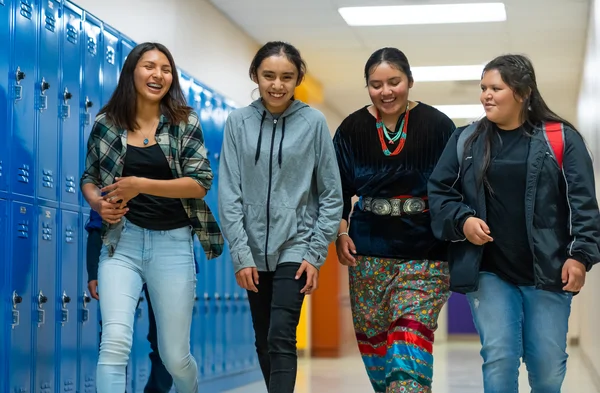 Four teenage girls walk and laugh together in a school hallway lined with blue lockers.