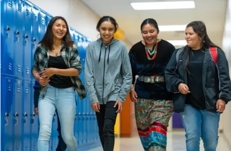 Four teenage girls walk and laugh together in a school hallway lined with blue lockers.