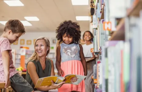 A woman reads to children in a library, surrounded by bookshelves and smiling kids.