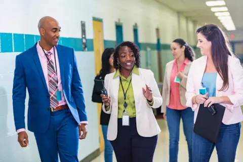 Five professionals walk and talk together in a brightly lit hallway, wearing business attire and carrying folders and badges.