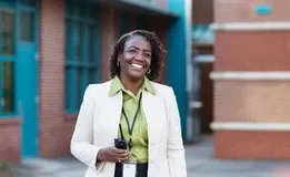 An educator, smiling confidently, stands outside of a school building with a cellphone in hand.