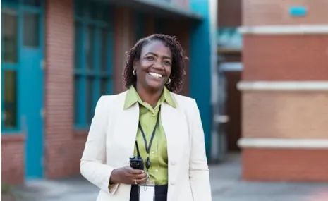 Smiling woman in a light blazer holding a walkie-talkie stands outside a brick building.