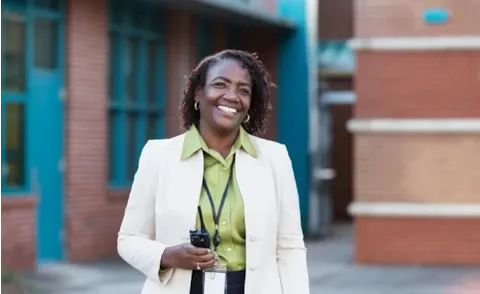 Smiling woman in a light blazer holding a walkie-talkie stands outside a brick building.
