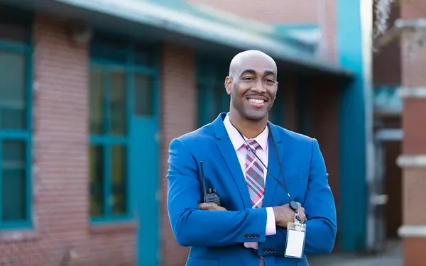 Smiling man in a blue suit with a lanyard stands confidently outside a brick building, arms crossed.