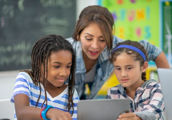 A teacher helps two children using a tablet in a colorful classroom with posters and a chalkboard in the background.