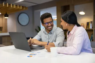 Two colleagues sit at a desk with a laptop, smiling and discussing documents in a modern office setting.