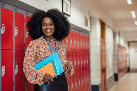 Smiling woman holding folders stands by red lockers in a school hallway.