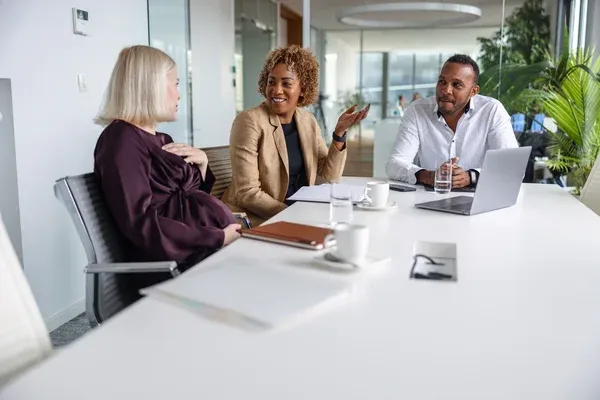 Three people sit at a conference table, talking and smiling, with a laptop and papers in front of them.