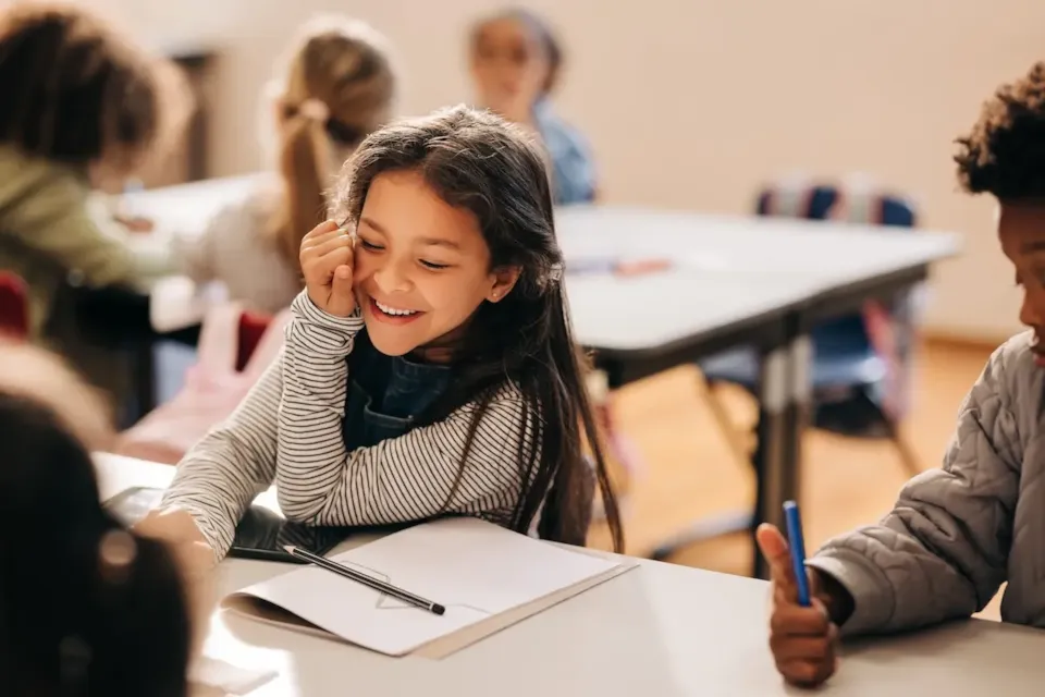 Smiling girl sitting at a desk in a classroom, writing in a notebook with classmates around her.