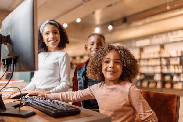 Three smiling children using a computer together in a library, with bookshelves in the background.
