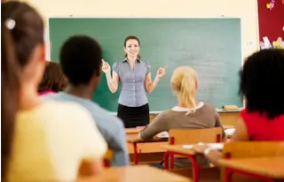 A teacher stands in front of a chalkboard, speaking to students seated at desks in a classroom.