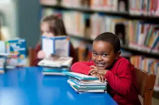 Smiling child in red shirt leans on a stack of books at a library table; shelves and another child in background.