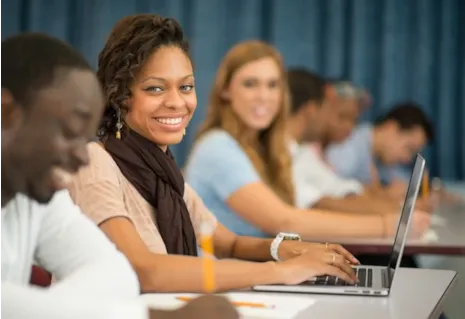 Young woman smiling at camera while using a laptop in a classroom with other students writing beside her.