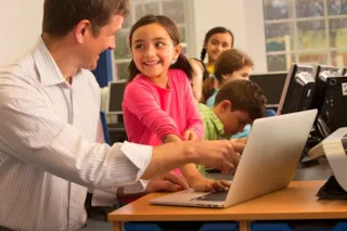 A teacher helps a smiling girl with a laptop in a classroom, surrounded by other students using computers.